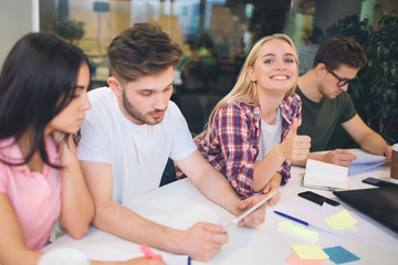 Picture of cheerful young blonde woman look on camer and smile. Other students work on their projects at table. They are serious and concentrated.