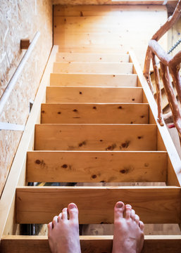 The legs of a man stand on the top step of a wooden staircase, in a house.