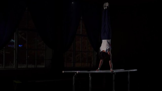 Male gymnast acrobat performs handstand on parallel bars in a dark room in slow motion sharing a somersault and landing on the floor. Training before the Olympic games