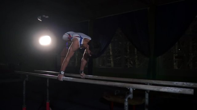 Professional gymnast in a dark room in sportswear stands on crayfish on parallel bars and performs flips in slow motion. Hard training in preparation for the Olympic games