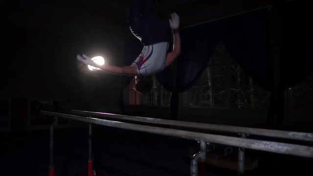 Male gymnast acrobat performs handstand on parallel bars in a dark room in slow motion sharing flips and landing on the floor