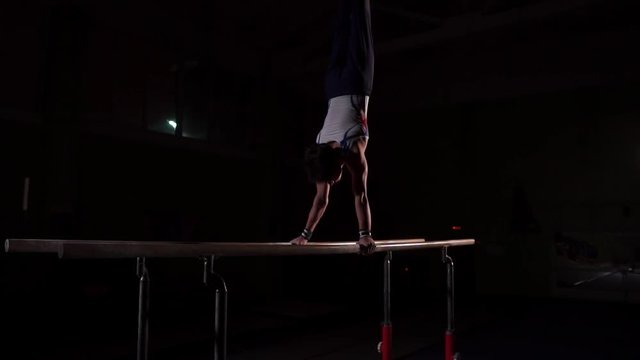 Professional gymnast in a dark room in sportswear stands on crayfish on parallel bars and performs flips in slow motion. Hard training in preparation for the Olympic games