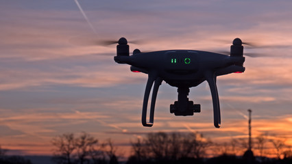 Naklejka na ściany i meble A drone hovers in flight in front of a sunset background. The drone is stationary hovering above the ground as the sunsets in the distance.