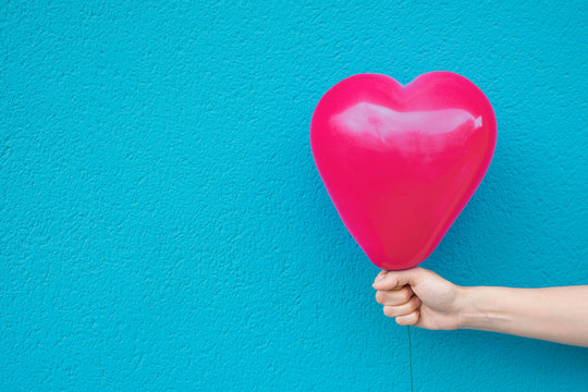 Young caucasian woman girl holds in stretched hand bright pink heart shape air balloon on turquoise painted wall background. Valentine love charity donation concept. Vivid Colors. Urban Atmosphere