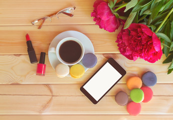 Coffee cup with colorful macaroons, smart phone, lipstck, nail polish and red peony flowers on wooden table. Top view, copy space.
