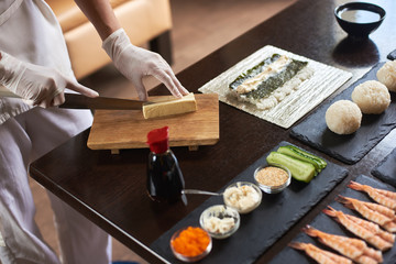 Close-up view of process of preparing rolling sushi. Hands in disposable gloves slicing omelet on wooden board
