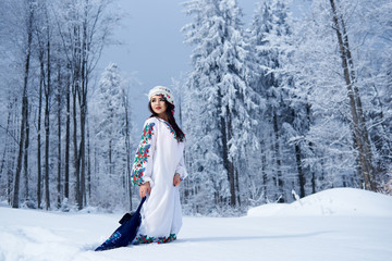 Fototapeta na wymiar Young attractive fashionable dark-haired woman in white dress embroidered with floral design, colorful kerchief, with blue linen shoulder bag in deep snow outdoors on spruce forest winter background.