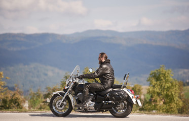 Side view of bearded motorcyclist in sunglasses and black leather clothing riding cruiser motorbike along narrow asphalt path on sunny summer day