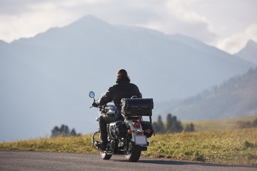 Back view of biker in black leather jacket riding motorcycle along road on blurred copy space background of beautiful foggy woody mountain range and cloudy sky on bright sunny summer day.