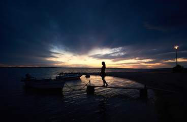 Fototapeta premium Silhouette of girl in short dress standing on narrow wooden bridge at small motor boats anchored at shore in sea inlet calm shallow water chained to pier on bright cloudy sky at sunset background.