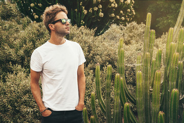 Young handsome bearded guy wearing a blank white t-shirt and sunglasses is standing in the garden...