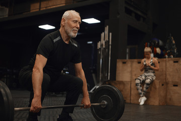 Fototapeta na wymiar People, age, sports, vitality and powerlifting. Picture of muscular fit mature retired man with strong arms exercising with barbell posing in gym with his young female instructor in background