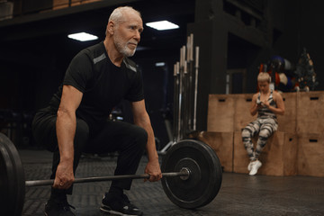 Fototapeta na wymiar Indoor shot of handsome unshaven male seventy year old male athlete in stylish sports clothes enjoying powerlifting training, holding barbell, doing squats, smiling confidently. Selective focus