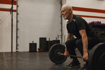Obraz na płótnie Canvas Stylish seventy year old male choosing healthy active lifestyle, training with weights indoors, lifting barbell, bulding muscular strong arms, having joufyl happy facial expression. Age and vitality