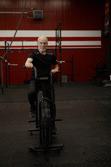 Fototapeta na wymiar Vertical image of healthy self determined energetic elderly man with gray beard sitting on stationary bike, having cardio workout in gym. Senior male cycling on bicycle, wearing sports clohtes