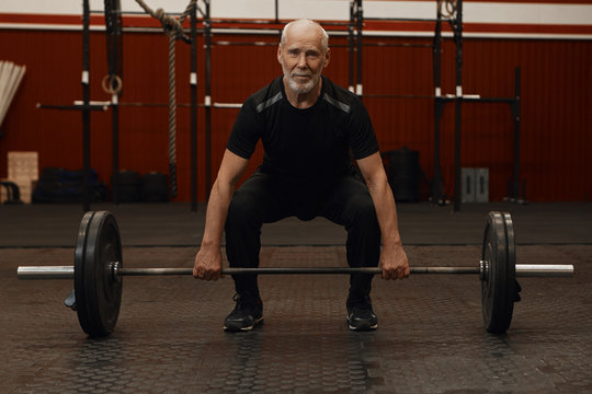 Attractive bearded retired male in black sportswear doing squats and lifting weights in gym. man smiling while working out, shaping up, squatting with barbell. Healthcare, motivation and retirement