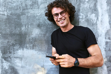 Portrait of smiling man standing outdoors texting on mobile phone. Young male with curly hair wears...