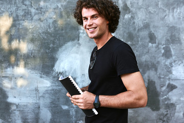 Outdoors image of happy man with curly hair, writing some notices on paper and preparing for exams, wearing black t-shirt posing on concrete wall. Freelancer businessman makes plan for a day