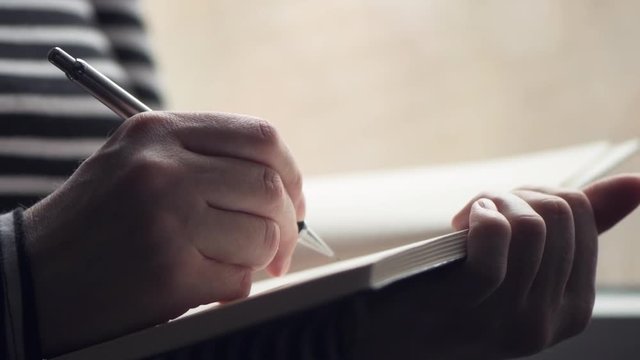 Woman writing diary by the window of her living room, close up with selective focus
