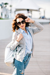 Pretty brunette girl with short hair is walking in city.  She wears shirt, jeans, jacket and bag. She touches sunglasses on face and looks to the camera.