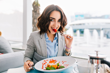 Portrait of pretty brunette girl with short hair at table  on terrace in restaurant. She has lunch time, eating healthy food, looking to camera.