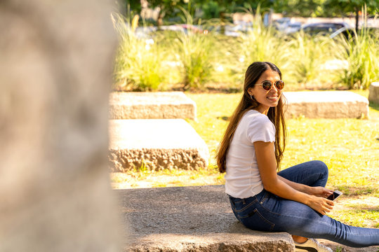 Portrait of a beautiful smiling young woman in a white shirt and sunglasses while sitting on a concrete block on a sunny day.