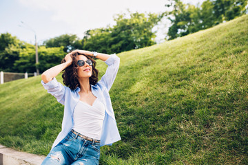 Attractive  brunette girl with short hair in sunglasses is posing to camera in park on meadow background. She wears white T-shirt, blue shirt, jeans. She keeps hands on head and looks happy.