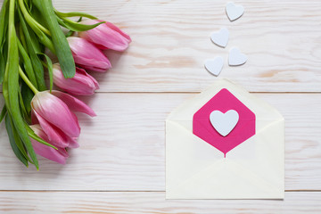 Obraz na płótnie Canvas Bouquet of pink tulips and hearts pattern in paper envelope. Top view, close-up, flat lay on white wooden background
