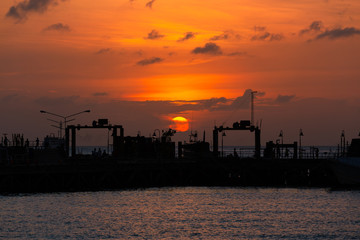 Fototapeta premium Suratthani, Thailand - January 7, 2019: Sunset in the sea at Nathon The ferry pier has tourists to sit on the pier. Big sun Beautiful orange sky on the January 7, 2018 in Samui Island, Suratthani, Tha