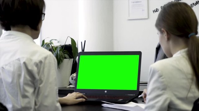 Young women working in office, sitting in chairs in front of computer with green screen, rear view. Two girls co workers looking at computer monitor with chroma key over white wall background.