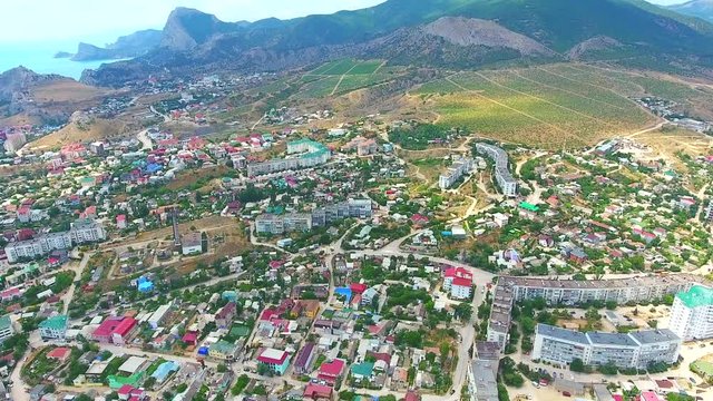 Drone footage of city resort Sudak coastline with green mountains, buildings and beach. Aerial: beautiful city centre, street and coastline Black sea, Crimea.