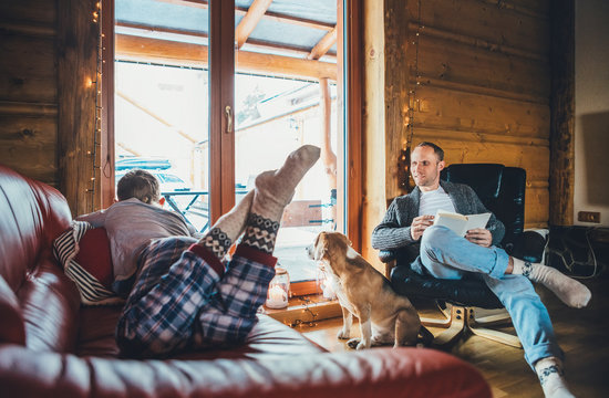 Father, son and his beagle dog spending holiday time in cozy country house. Dad reading a book, boy lying and listening, dog looking in window. Winter family vacations concept image.