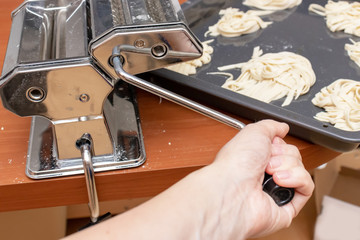 Hand holding a handle of rolling dough machine fo making pasta and ravioli close up with homemade ribbon egg italian pasta on background