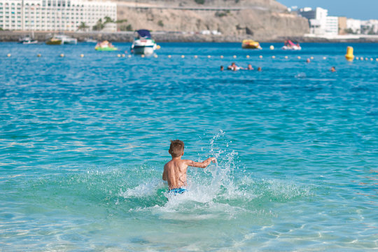 Cute European boy in colorful swimming shorts is running towards the ocean, making numerous splashes around. He is happy to spend his summer holidays at the sea cost.