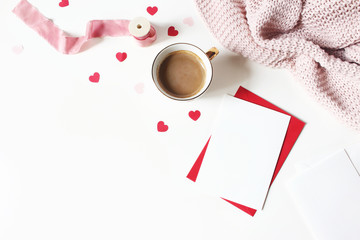 Valentines day or wedding still life scene. Cup of coffee, envelope, paper hearts confetti, knitted plaid and blank greeting card mockup on white table background. Love concept. Flat lay, top view.