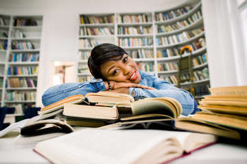 Portrait of positive african american girl putting her arms in to the stack of books and looking at camera with smile