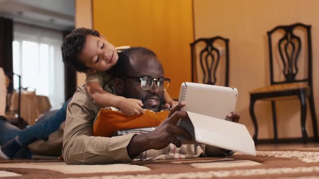 Tilt up shot of biracial boy laying on his father's back and watching over dad's shoulder on tablet computer's screen