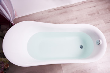 Top view of a white bath tub filled with clear water standing on a light brown wooden bathroom floor