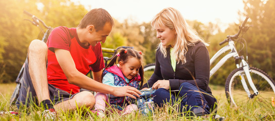 Fototapeta premium Young happy family tourists bikers, mom, dad and child resting with bicycles on grassy hill, Healthy lifestyle and relations concept.