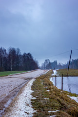 Obraz premium Empty Countryside Landscape in Cloudy Winter Day with Snow Partly Covering the Ground and Fog, Puddles and Tire Marks on the Road