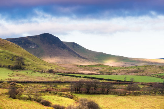 Dramatic purple-blue clouds over a sun-dappled landscape of green fields, hills, and mountains in Glenariff, County Antrim, Northern Ireland near Cushendall
