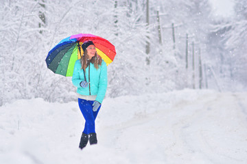 Happy lonely girl with rainbow umbrella on a rustic empty road. Snowy winter weather.