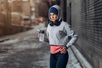 Fit woman drinking protein shake on winter day in city. Sporty woman in headphones