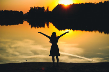 Silhouette of a young beautiful girl with hands up against the background of the sunset in the reflection of the pond