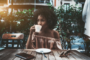 Thoughtful charming curly African-American girl is drinking delicious coffee from a white ceramic cup and pensively looking aside while sitting alone in a street bar on a sunny summer day