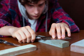 young man sharpening a knive on wooden table on dark background b