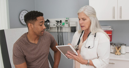Portrait of young black man consulting with mature female doctor indoors medical clinic. Close up of senior caucasian doctor using tablet computer to share test results with male black patient