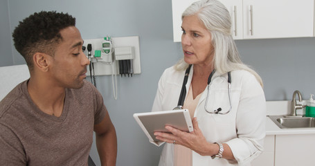 Mature white female doctor using portable computer pad to take patient notes. Young male...