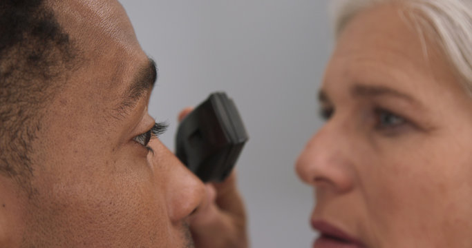 Young Black Male Patient Having His Eyes Checked By Senior Female Doctor. Mature Female Doctor Using Opthalmoscope To Examine Eyes Of African-american Patient