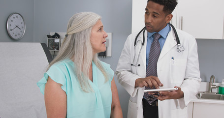 Male millennial MD taking notes on electronic tablet while listening to senior female patient....